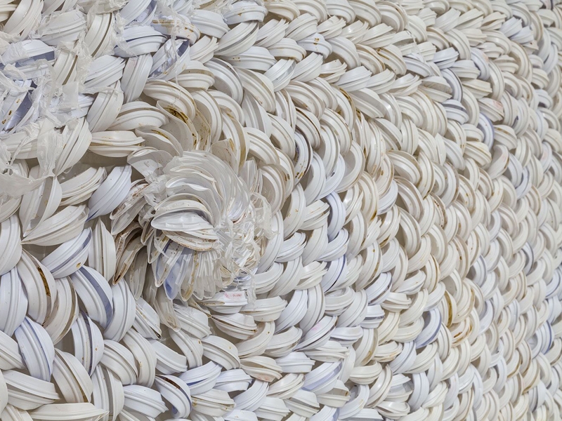 Sweeping and flowing used coffee lids create this woven detail of Susie Ganch’s Pile: Triangle Trade, 2015, shown at Mixed Greens, photo courtesy of Mixed Greens Susie Ganch, Pile: Triangle Trade