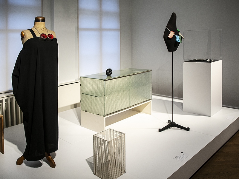 Exhibition view, Jewellery & Garment. The Bröhan’s "Minimalism Room" is furnished with Gerrit Thomas Rietveld’s sideboard made of industrial glass (shown in the background). On it rests David Bielander’s Pneu bangle, 2010, blackened silver in the shape of a tire. Other works on display, (left mannequin) jewelry: Daniel Kruger: Untitled, 2006, necklace, silver, pigment. Garment: Yohji Yamamoto, dress, 2014, wool. (Between the mannequins) Josef Hoffmann: white lacquered lattice object, Wiener Werkstätte. (Right mannequin) Lisa Walker, Untitled, 2019, necklace, mobile phones, thread